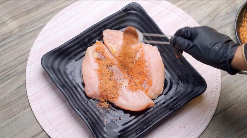 Season the chicken Raw chicken pieces being coated with Cajun seasoning, with a few herbs and spices scattered around the cutting board.