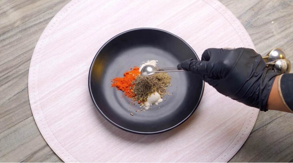 Cajun seasoning A small glass bowl filled with vibrant red-orange Cajun seasoning, surrounded by scattered whole spices like peppercorns, dried chili flakes, and a dusting of paprika on a rustic wooden surface.
